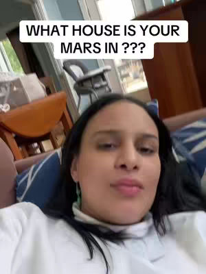 A woman with dark hair is shown in a close-up shot, positioned against a background of a room with a window and furniture. She is looking directly at the camera. A rectangular text overlay at the top reads 'WHAT HOUSE IS YOUR MARS IN ???'.
