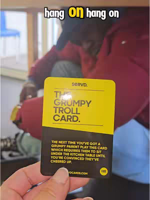 A hand holds up a brightly colored card with text on it, while the blurred background suggests someone is sitting under a table. The card is primarily yellow and black with the words, "The Grumpy Troll Card." written on it.