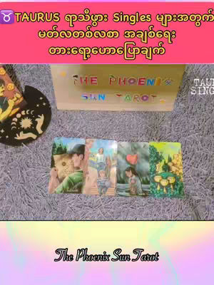 The image shows a collection of tarot cards laid out on a surface that appears to be carpet, and a sign that says the the phenix sun tarot. Above these elements, there is a pale pink banner containing text in a non-english language, likely Burmese. The bottom of the image has the text "The Phoenix Sun Tarot" in an elegant font.