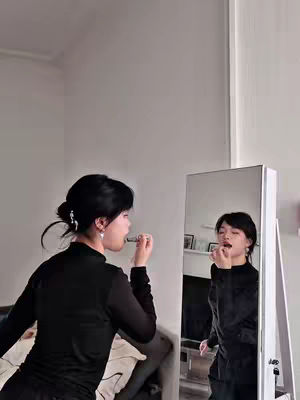 A woman in a black top applies lipstick while looking in a mirror. Her reflection is also visible, and the background is a simple room with a wall and a glimpse of furniture.