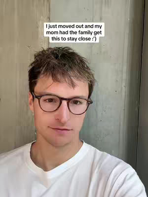 A young man with short brown hair and glasses stands in front of a concrete wall. A text overlay is positioned above the man's head. He is wearing a white t-shirt. The lighting is natural and the man is looking directly at the camera.