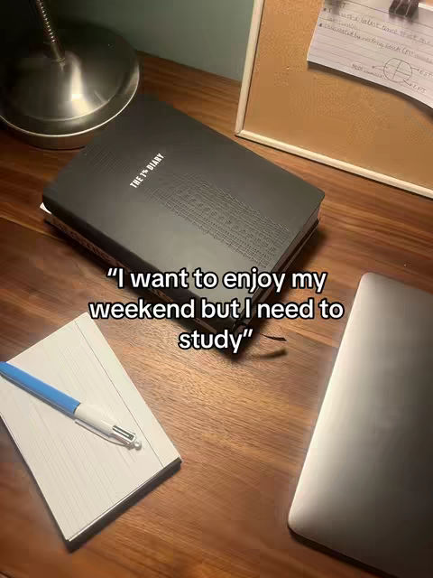 An overhead shot of a wooden desk with a black diary, a notebook with a pen, and a laptop. Text overlayed at the bottom reads "I want to enjoy my weekend but I need to study".