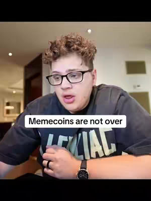 A young man with curly brown hair and glasses is looking at the camera with a slight open mouth. He is wearing a black t-shirt and a watch on his wrist. Text in a white bar at the bottom reads "Memecoins are not over". The background is a brightly lit interior.