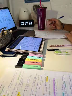 A well-lit desk with a laptop, tablet, papers, highlighters, and a digital clock displaying the time. A person's hand is visible, holding a pen and working on the papers. A purple tumbler with a straw, a cat-shaped lamp, and other stationery items are on the desk, creating a study environment.