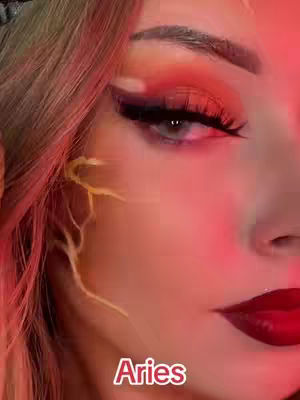 A close-up shot features a woman's face with striking makeup. Her eye is heavily made up with vibrant orange and red eyeshadow, dramatic black eyeliner, and long, dark eyelashes. The background is softly blurred with a pinkish-red glow. The text 'Aries' is superimposed at the bottom.