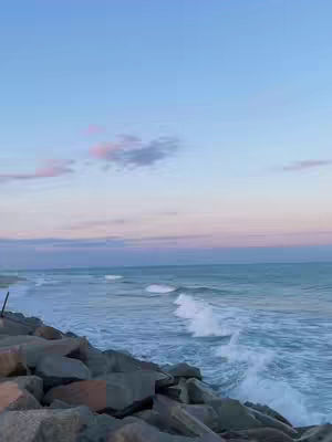The video cover image captures a scenic coastal view at dawn or dusk. The sky transitions from a soft blue at the top to hues of pink and purple towards the horizon line, where the ocean meets the sky. Waves are rolling toward the shore with the tide coming up against rocks in the foreground. The composition is framed vertically, emphasizing the vastness of the sky and the expanse of the sea.