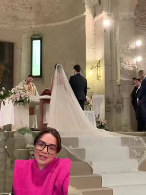 A woman with glasses is centered in the foreground, smiling and facing the camera. Behind her, a wedding ceremony is taking place in a stone building with arched windows and white walls, the bride and groom are standing at the altar.