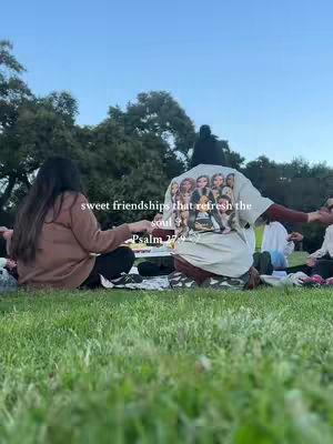 Two people are sitting on a picnic blanket in the grass, facing each other with other people in blurred background. One person wears a white t-shirt with faces on it and has their back to the camera. Text overlays the image with a religious quote.