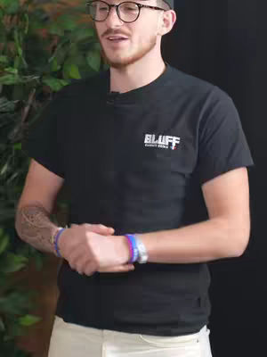 A man wearing glasses, a black t-shirt with the word "BLUFF" and a logo is standing in front of a blurred background. He is wearing a black cap and has visible tattoos on his arm. He is looking up as if he is speaking.