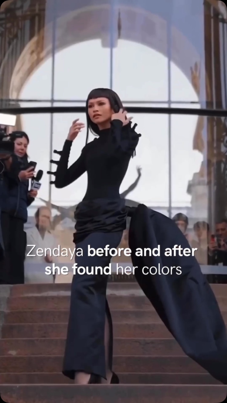 Zendaya in a stylish black outfit descends a set of stairs, posing for photos. She wears a sleek black top, a draped skirt, and her hair is styled with a short, blunt fringe. The background shows a modern architectural setting, likely an event or fashion show, with other people taking pictures.