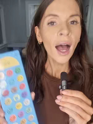 A woman with brown hair is holding a communication board. She has her mouth open as if speaking into a microphone.