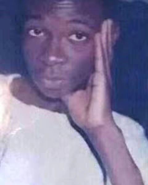 The cover image shows a medium close-up of a dark-skinned individual, likely a man, resting his hand near his face. The person is looking directly at the viewer with a serious expression. The background is a blurry shade of dark blue or purple, which contrasts with the lighter tones of the subject's skin and clothing, the person seems to be wearing a white shirt.