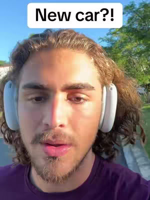 A man with a beard and curly brown hair, wearing a purple shirt, with white headphones. The background shows a bright sky and some greenery.