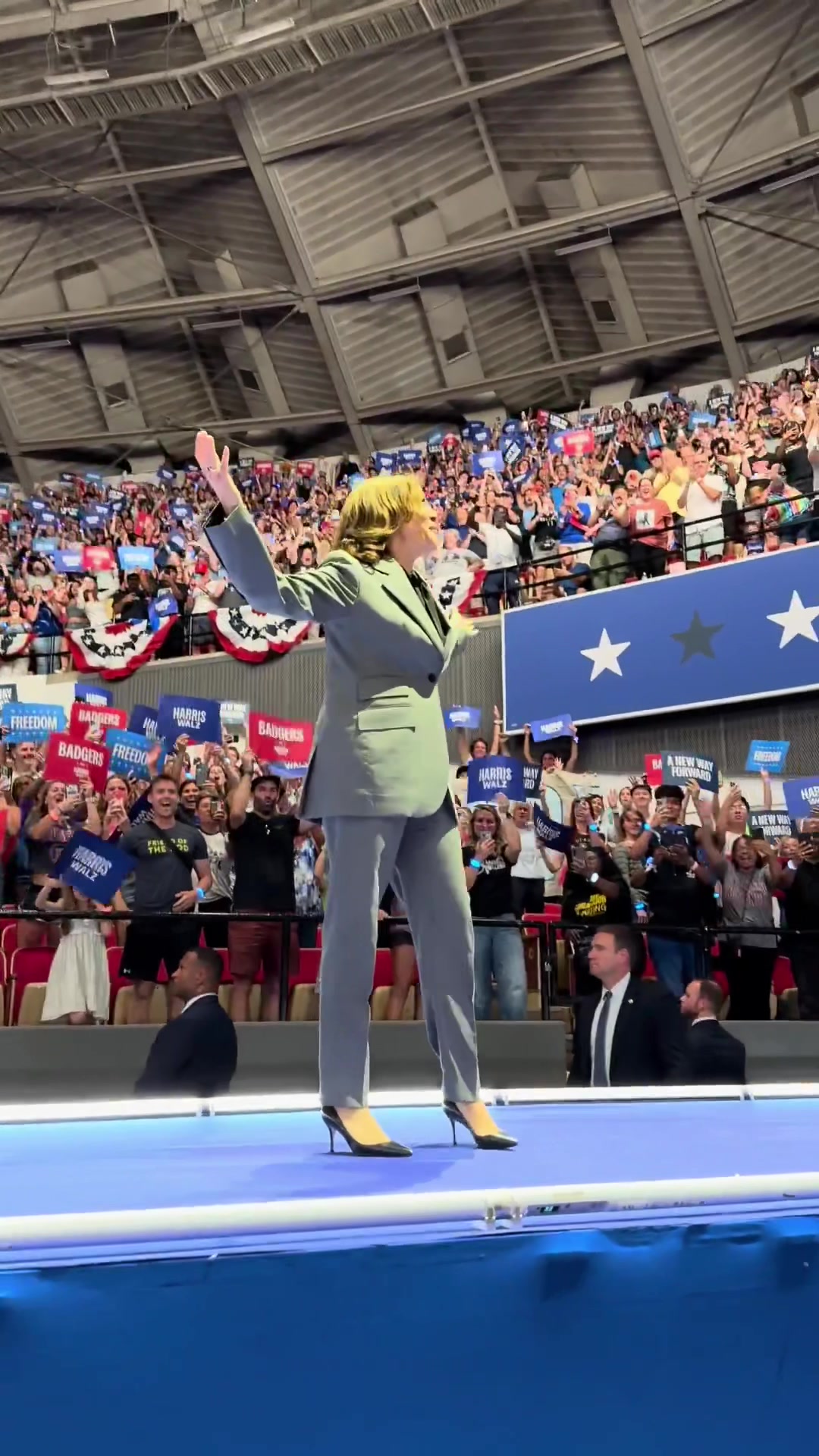 A woman in a grey suit with arms raised stands on a stage, addressing a crowd of people. The stage is illuminated by bright lights. The background is filled with a large audience, holding signs and cheering. The overall composition is a dynamic representation of a political rally or speech.