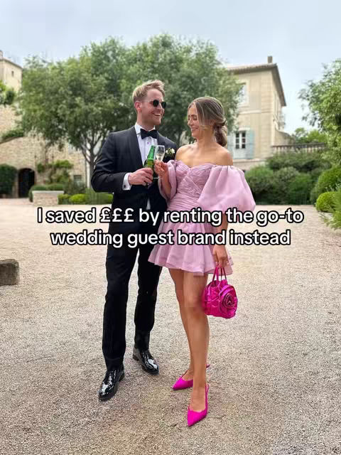 A couple is standing outdoors, posing for a photo. The woman is wearing a short, pink dress and holding a pink purse. The man is in a black suit and sunglasses. The background suggests a garden setting with buildings.