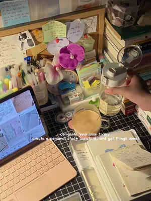 A desk setup with a laptop, notebooks, and various stationery items. A hand holds a water bottle. The background includes a bulletin board covered with notes and decorative items