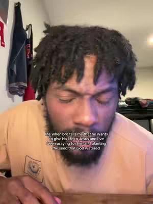 The image shows a Black man in a close-up shot, looking down with eyes closed and a slightly furrowed brow, suggesting deep thought or prayer. He has short, dark, curly hair and a beard. He is wearing a tan-colored shirt. Text overlaid on the image indicates that he is reacting to a friend wanting to commit his life to Jesus, conveying a message of faith and spirituality. The background is blurred, featuring parts of a wall with some clothes hanging, providing a home setting.