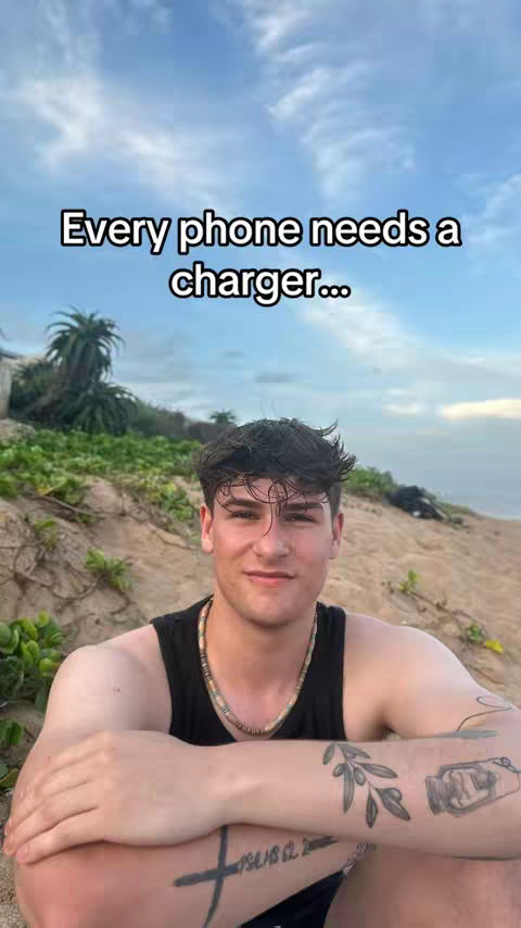 A young man is the central figure, with a beach background and text overlay. He's wearing a black tank top and a necklace, with arm tattoos. The text reads, 'Every phone needs a charger...' against a backdrop of a cloudy sky, ocean, and some greenery.