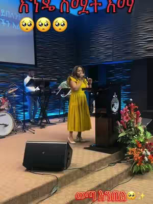 A woman in a yellow dress is singing into a microphone on a stage during a church service. There is a screen behind her, and a drummer and keyboard players. The stage is decorated with flowers.