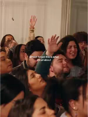 A crowd of people, primarily young adults, are gathered closely together, many with their faces tilted upwards and expressions of joy or reverence. Arms are raised in the air. Warm lighting suggests an indoor setting, possibly a church or community gathering.