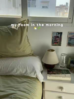A bedroom scene featuring a bed with a white top sheet and olive-green comforter, a pillow, a bedside table with a lamp (red base, beige shade), a glass of water, and a picture on the wall. The room is bright with natural light coming from a large window.
