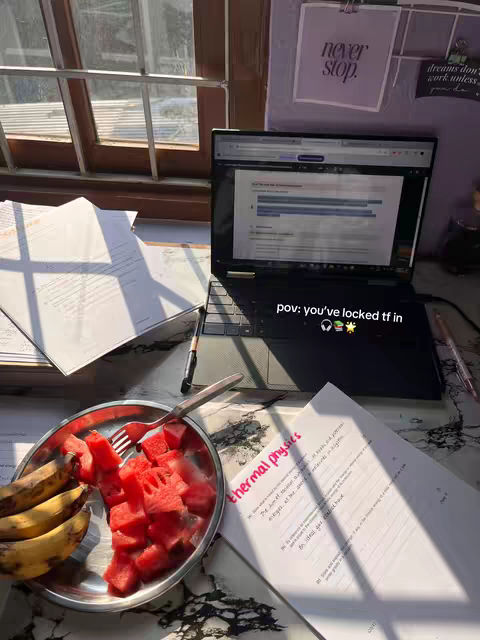 A desk setup with a laptop, papers, and a plate of snacks. The laptop screen displays text, a plate of watermelon and bananas sit nearby, and a window is visible in the background. The lighting suggests a natural light setting.