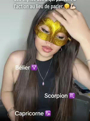 A woman with long brown hair wearing a gold mask covering her eyes and a black top is facing the camera. Overlay text including astrological signs and their symbols are added to the image. The background is a blurred room with neutral colors. The woman is holding her right hand to her head while smiling softly with the mask on.