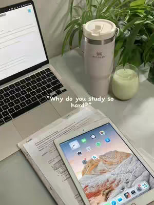 A workspace featuring a laptop with an open screen, a tablet displaying a desert landscape, a stainless-steel tumbler, and a glass with a lime green substance in it, set against a background of green plants.