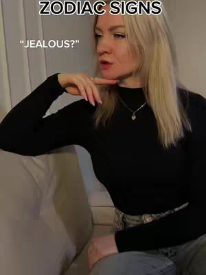 A woman with blonde hair is seated on a gray sofa, her left arm resting on her lap and her right hand touching her chin. She is wearing a black long-sleeved shirt and jeans. Text overlays at the top read "ZODIAC SIGNS", and "JEALOUS?" in the upper left corner.