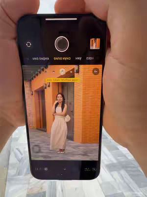 A person holding a phone is showing the camera view. The phone displays a woman in a cream-colored dress posing in front of a building with orange walls. A yellow box with text is on the screen.
