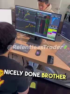 A top-down shot shows two men in a home office setting. The foreground features a person facing away from the camera, wearing eyeglasses and a black t-shirt with a yellow design. The background holds a large computer monitor displaying stock trading charts and data, with another person visible behind the monitor. A desk is positioned with a mouse, keyboard, and smartphone. The overlay text is yellow on a dark background.