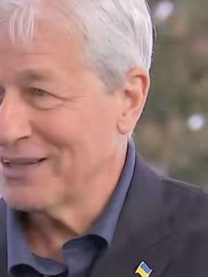 A close-up shot features a man with gray hair, a dark blue shirt, and a dark blazer. The background is blurred. A small pin with the Ukrainian flag is visible on the blazer. The man is looking slightly to the side with a subtle smile.