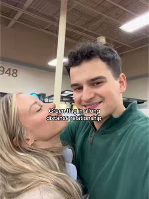 A man and woman are shown in a close embrace. The man, with dark hair and a green shirt, is smiling towards the camera. The woman, with blonde hair, is leaning in to kiss the man. Text overlayed reads, "Green flag in a long distance relationship."
