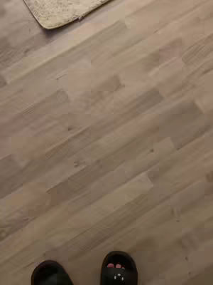 The image captures a first-person perspective of a person's feet clad in black slides with light-colored wooden flooring in the background. A beige rug is partially visible. The lighting appears natural, and the composition is simple, emphasizing the floor and footwear.