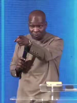 A man in a brown shirt is holding a microphone and pointing. The background is a solid blue.