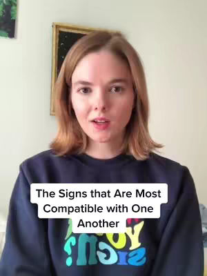 A woman with short, light brown hair is centered in the frame, speaking directly to the viewer. She's wearing a dark blue sweatshirt, and a text overlay at the bottom of the screen reads, 'The Signs that Are Most Compatible with One Another.' A framed picture hangs on the wall behind her, adding a hint of decor to the simple indoor setting.