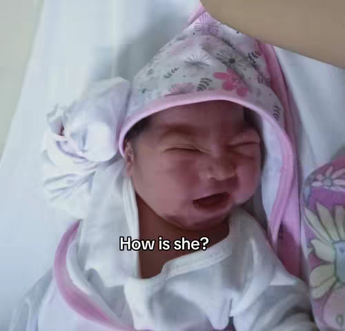 The image features a newborn baby wrapped in a white blanket and wearing a pink-trimmed bonnet. The baby has a slightly grumpy expression, with a furrowed brow and a slightly open mouth as if crying. The text overlay 'How is she?' is present in a white font at the bottom of the image.
