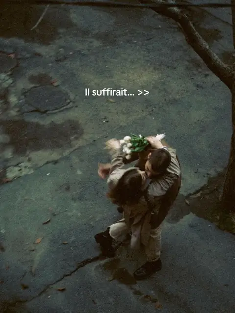 Overhead shot of a couple embracing outdoors, possibly in a courtyard. The ground is wet, and the scene is muted with cool tones. The couple is holding a bouquet of white flowers.