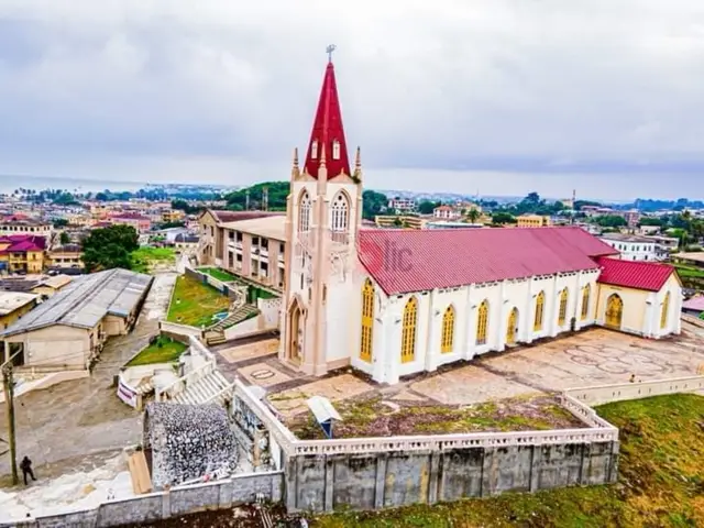 An elevated, wide-angle shot showcases a cathedral with a red-tipped steeple, a white facade, and a red roof, set against a backdrop of a cloudy sky and surrounding urban landscape. The church is the focal point, positioned centrally with a clear view of its architectural details. Green foliage and urban structures encircle the cathedral, while a low, light-colored wall and a person are at the bottom left foreground.