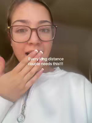 A woman with fair skin and glasses looks at the camera with a surprised expression, her hand covering her mouth. Text overlay at the bottom says 'Every long distance couple needs this!!!'. The background appears to be an indoor setting.