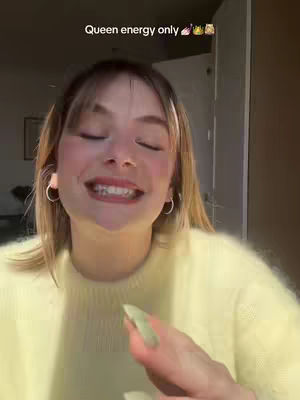 A young woman with fair skin and light-colored hair smiles widely with her eyes closed. She wears a yellow sweater and silver hoop earrings. The background is blurred, with a suggestion of an interior space with a door and a window. A text overlay at the top says 'Queen energy only' followed by emoji.