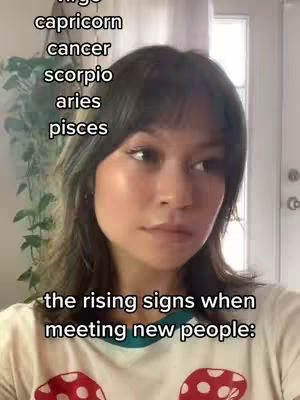 A selfie-style image of a woman from the chest up. She is looking slightly to the side with a neutral expression. The background is blurred but appears to be a room with window and possibly a plant. Text overlays list zodiac signs, and another overlay states "the rising signs when meeting new people:"