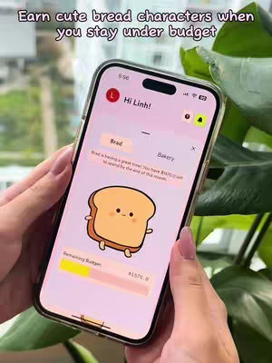 The cover image shows a smartphone held by a person with well-manicured nails, likely a female. The phone screen displays an app interface with a light pink background. Text overlays at the top say, "Earn cute bread characters when you stay under budget". Inside the app interface, it reads "Hi Linh!" with a pink bread character, and the bottom of the screen has a yellow and pink bar. Behind the phone, there's a soft-focus background of green leaves, indicating a visually appealing presentation.