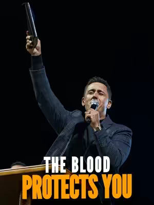 A man in a suit, holds a microphone in one hand and raises an object above his head with the other. Text overlayed on the bottom highlights the phrase 'THE BLOOD PROTECTS YOU'. The background is dark and blurry.