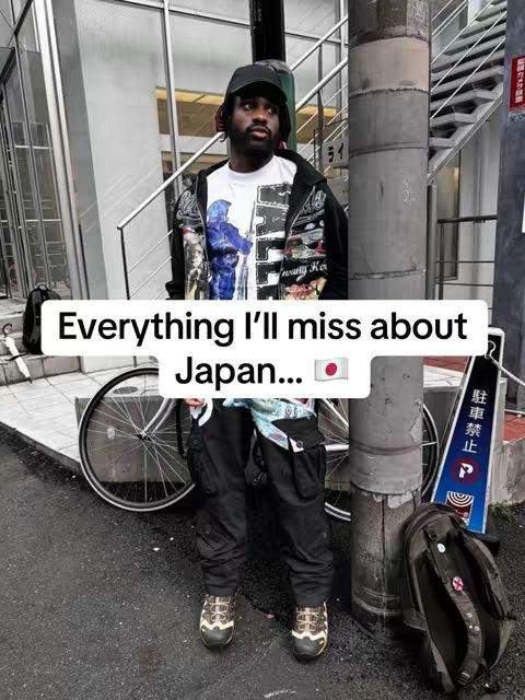 A man stands on a city street, posing in front of a bicycle and a large concrete pillar. He is wearing a black cap, a jacket, and black pants. Text overlay on image reads 'Everything I'll miss about Japan...'