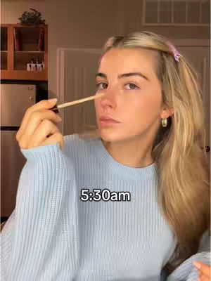 A woman holds a makeup applicator to her nose while looking at the camera. The background has a kitchen with a refrigerator and wooden cabinets. The woman is wearing a light blue sweater and her hair is long and blonde, parted to the side.