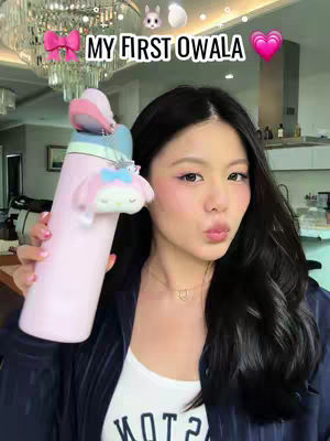 A woman with long black hair holds a pink water bottle with a My Melody charm. The background suggests a well-lit indoor setting with a chandelier.
