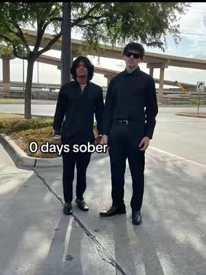 Two men stand side by side on a sidewalk, dressed in black. The background features a blurred cityscape with bridges and trees. A text overlay says '0 days sober'.