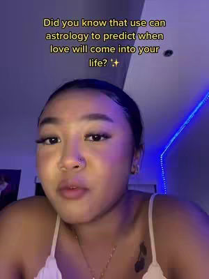 A selfie-style shot featuring a young woman with dark hair and a fair complexion. She is positioned slightly off-center, looking up and to the side with a somewhat inquisitive expression. The background is dimly lit and shows an indication of a blue neon light. Overlaid on the top is bright yellow text that poses a question about astrology and love, and there is sparkle symbol. She has a small nose ring and appears to be wearing light makeup and a white strap top. The lighting is soft, casting subtle shadows, and the overall feel is casual and approachable.