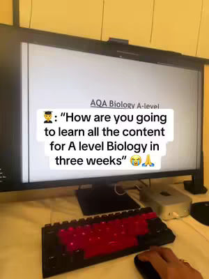 The cover image shows a computer monitor displaying a white document with the text "AQA Biology A-level." Overlayed on the screen is a text bubble with the question "How are you going to learn all the content for A level Biology in three weeks" with a graduation cap and prayer hands emoji. Below the screen is a black keyboard with red keys, the bottom right has a visible hand holding a computer mouse. The background is a desk and the wall behind the monitor.